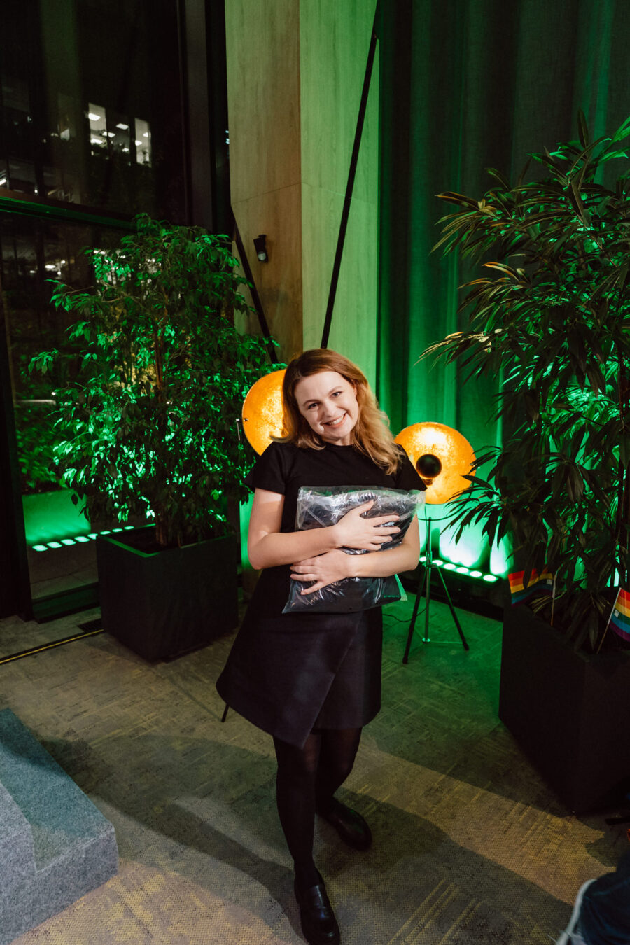 zdjecia eventowe aula polska bnp paribas warszawa 097 Uśmiechnięta kobieta w czarnym stroju stoi w pomieszczeniu, przytulając poduszkę. Jest otoczona zielonymi roślinami i ciepłymi żółtymi światłami, z dużymi oknami i ciemnymi zasłonami w tle.