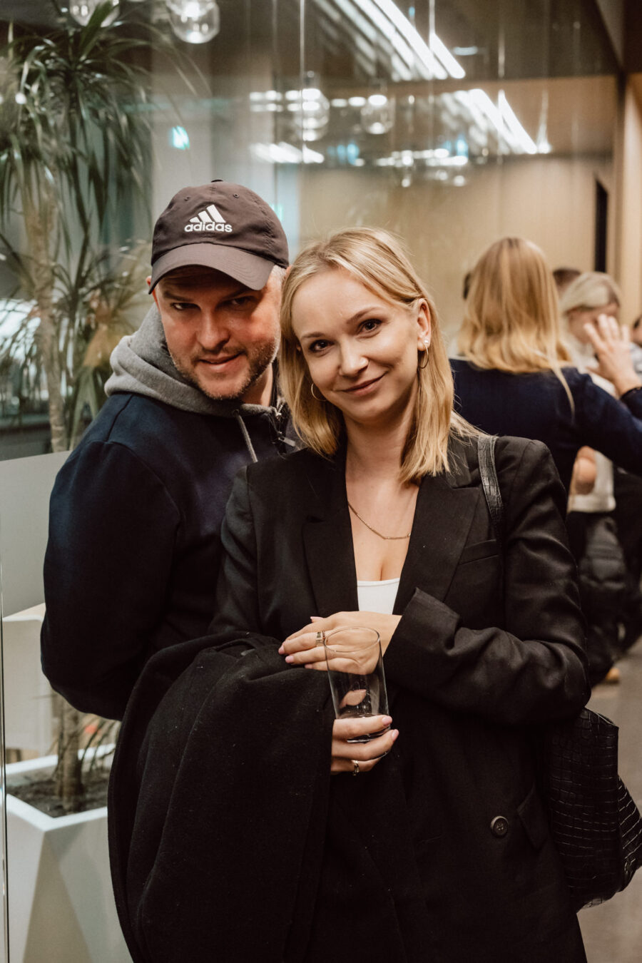 zdjecia eventowe aula polska bnp paribas warszawa 098 Mężczyzna w czarnej czapce Adidas i bluzie z kapturem stoi obok uśmiechniętej kobiety w czarnej marynarce trzymającej szklankę, w pomieszczeniu na imprezie towarzyskiej z innymi ludźmi i roślinami w tle.