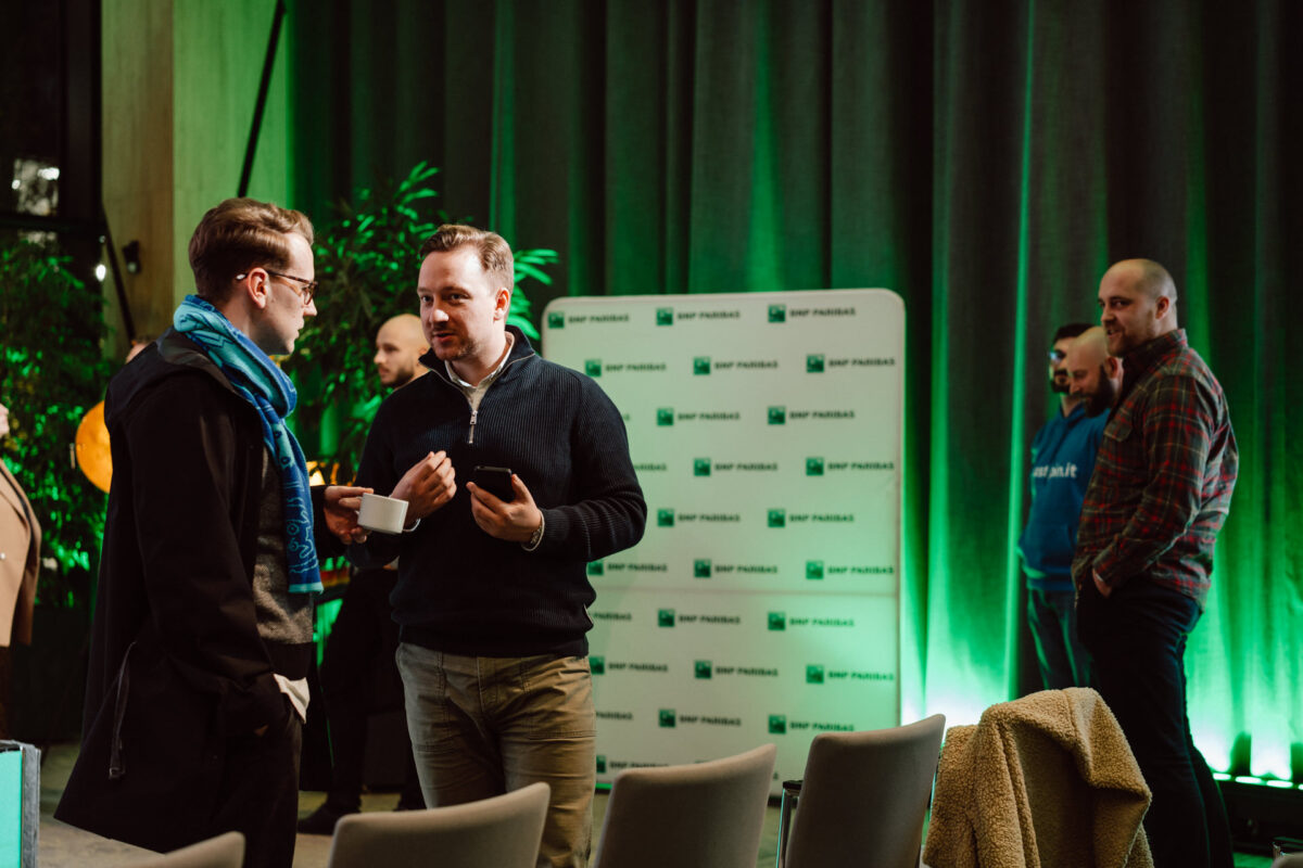 zdjecia eventowe aula polska bnp paribas warszawa 100 Dwóch mężczyzn rozmawia na pierwszym planie, jeden trzyma filiżankę i spodek, podczas gdy trzech innych stoi i rozmawia w tle. Za nimi widoczna jest zielona kurtyna i tło z marką BNP Paribas.