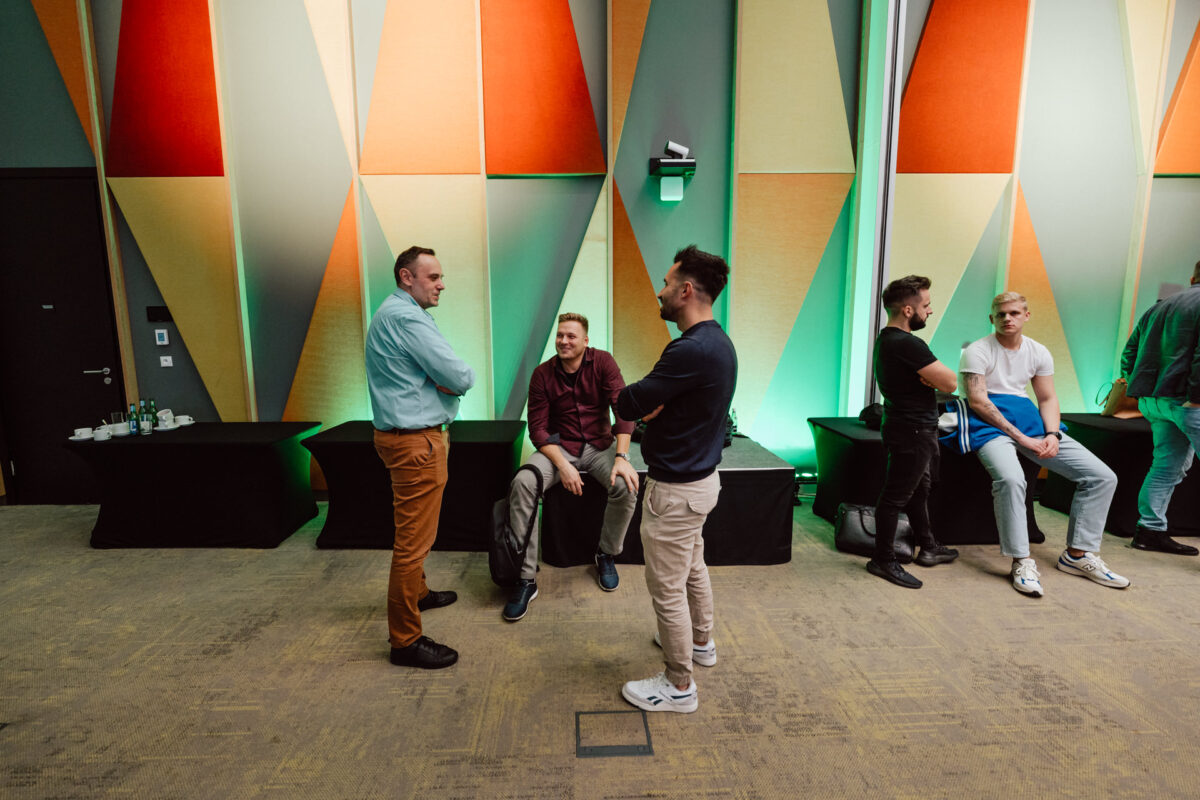 zdjecia eventowe aula polska bnp paribas warszawa 101 Kilku mężczyzn rozmawia i siedzi przy kolorowej geometrycznej ścianie w nowoczesnym pokoju z zielonym oświetleniem akcentującym. Trzech mężczyzn stoi i rozmawia, podczas gdy trzech innych siedzi na stołach wzdłuż ściany.