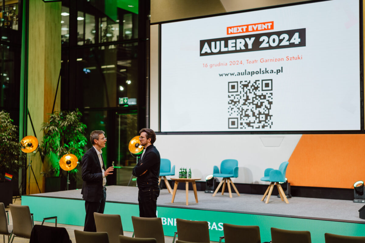 zdjecia eventowe aula polska bnp paribas warszawa 102 Dwóch mężczyzn stoi i rozmawia przed sceną z pustymi krzesłami. Duży ekran za nimi wyświetla "Next Event Aulery 2024" z kodem QR i szczegółami wydarzenia w języku polskim. Miejsce wydarzenia zdobią jasne światła i rośliny.