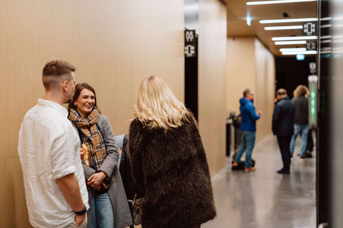 zdjecia eventowe aula polska bnp paribas warszawa 103 Trzy osoby stoją i rozmawiają w nowoczesnym korytarzu z jasnymi ścianami i jasnymi lampami sufitowymi. W tle widać dwóch mężczyzn i kobietę, którzy również rozmawiają w pobliżu czarnego znaku z białym napisem.