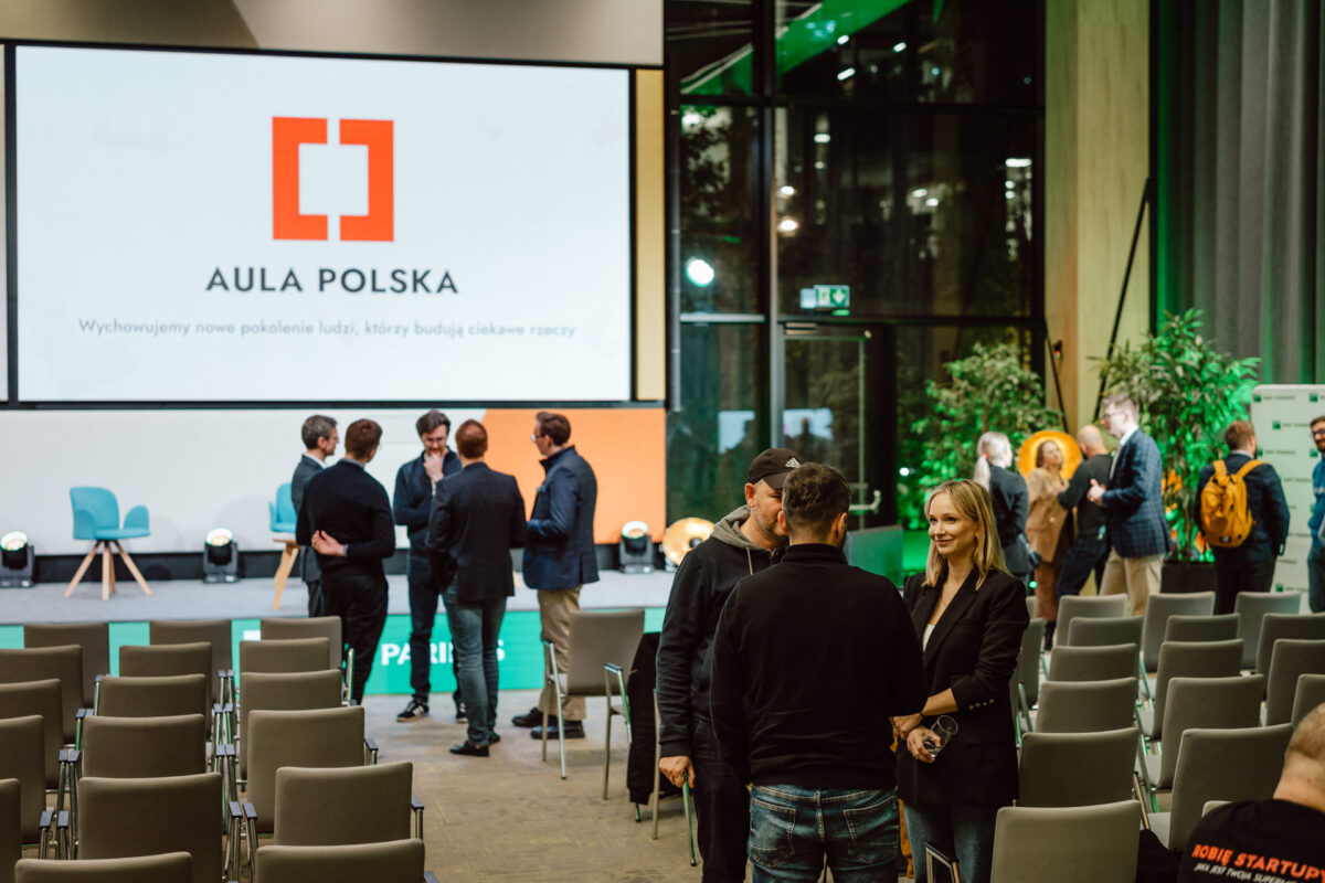 zdjecia eventowe aula polska bnp paribas warszawa 105 Grupa ludzi rozmawia w nowoczesnej sali konferencyjnej z rzędami krzeseł. Na dużym ekranie wyświetlany jest napis "Aula Polska" i slogan w języku polskim. Niektóre osoby stoją w pobliżu sceny, a inne rozmawiają w przejściu.