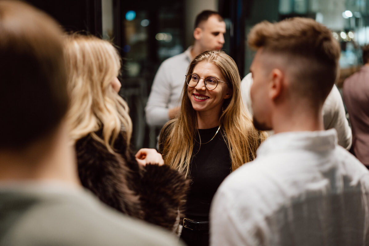 zdjecia eventowe aula polska bnp paribas warszawa 106 Kobieta z długimi blond włosami i w okularach uśmiecha się podczas rozmowy z małą grupą ludzi na spotkaniu towarzyskim w pomieszczeniu.