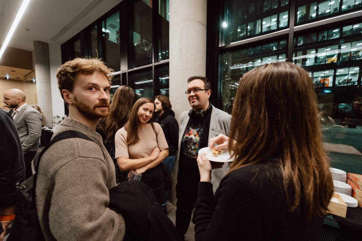 zdjecia eventowe aula polska bnp paribas warszawa 107 Grupa ludzi stoi i rozmawia podczas nocnego spotkania towarzyskiego w pobliżu dużych okien. Jedna osoba trzyma talerz z jedzeniem, podczas gdy inni są zaangażowani w rozmowę, a na zewnątrz widać światła miasta.