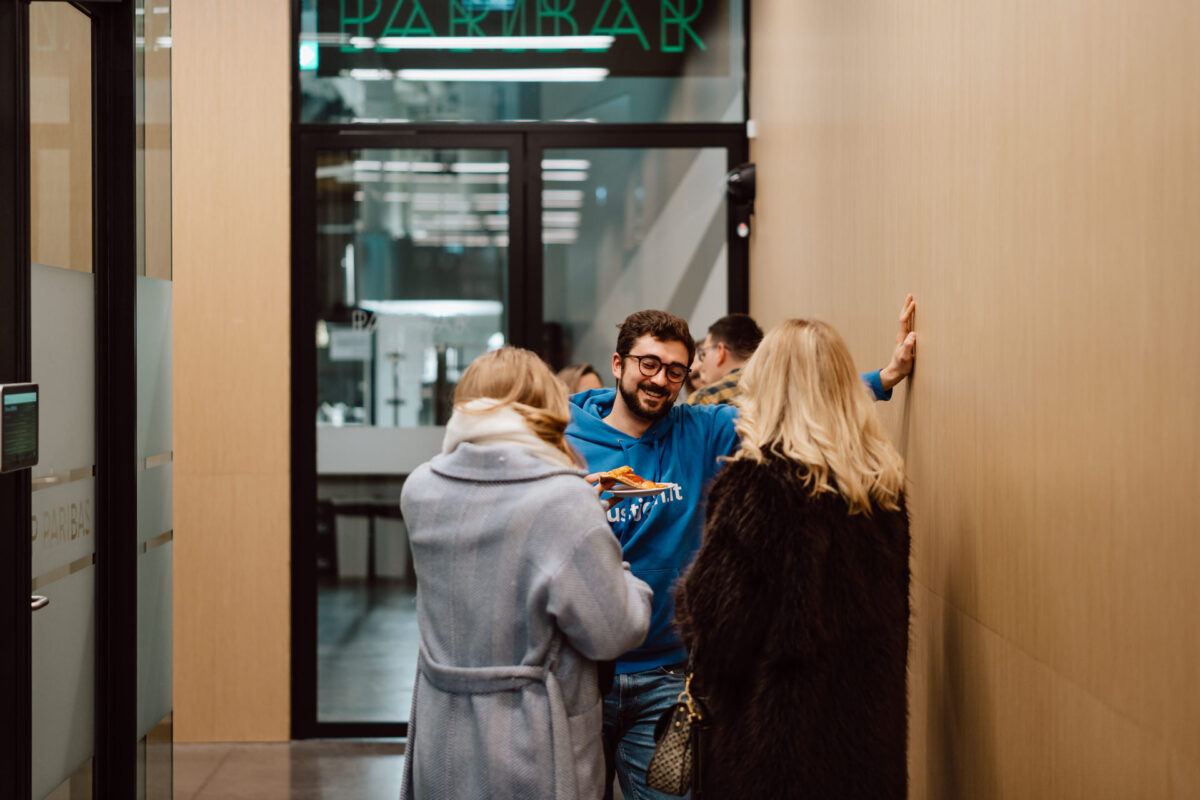 zdjecia eventowe aula polska bnp paribas warszawa 108 Trzy osoby stoją i rozmawiają w korytarzu; jeden mężczyzna w okularach i niebieskiej bluzie z kapturem uśmiecha się podczas interakcji z dwiema kobietami w zimowych płaszczach, z których jedna trzyma talerz z jedzeniem. W tle widać szklane drzwi i neon.