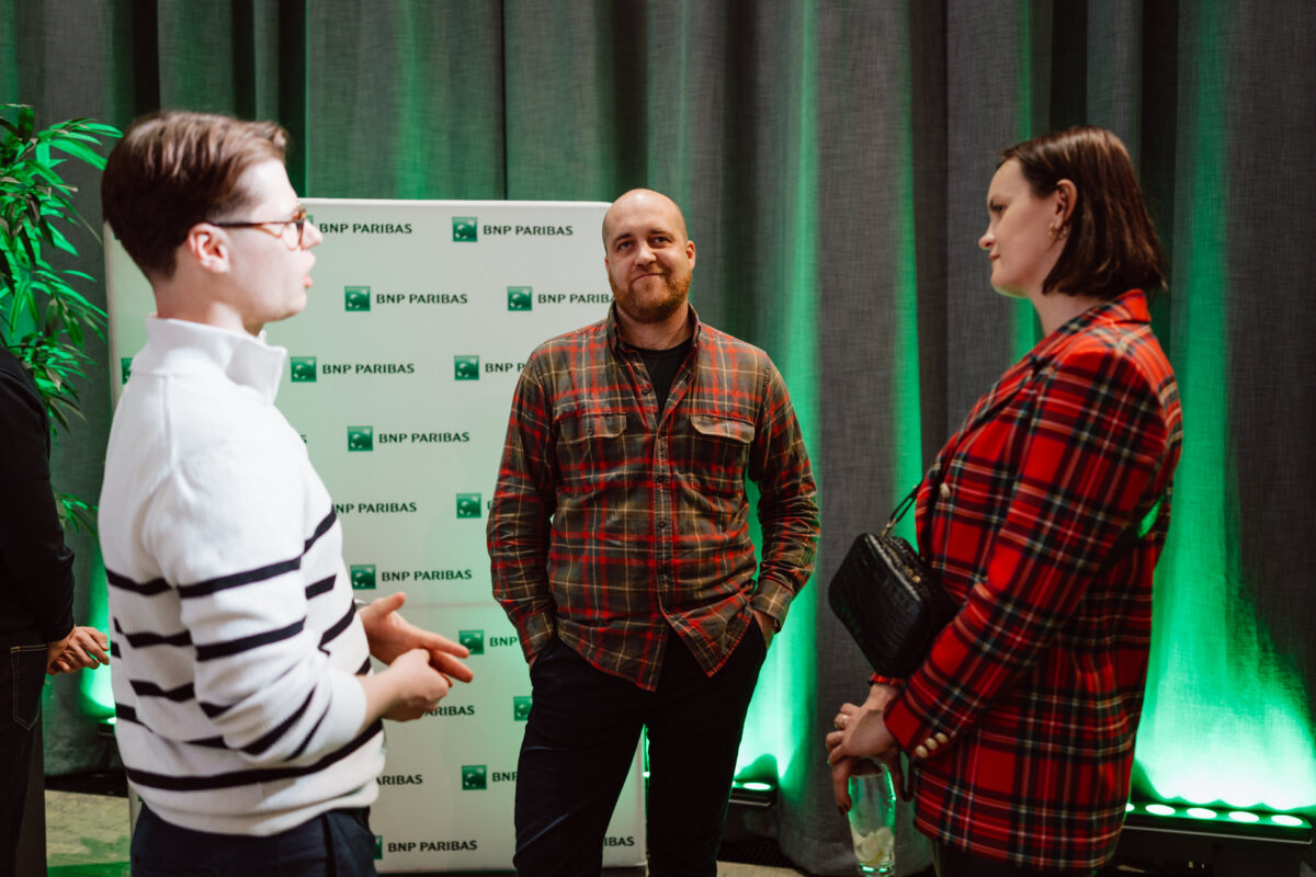 zdjecia eventowe aula polska bnp paribas warszawa 113 Trzy osoby stoją i rozmawiają na imprezie wewnętrznej z tłem marki BNP Paribas. Dwie osoby ubrane są w koszule w kratę, a jedna w biały sweter w paski. W tle widoczne jest zielone oświetlenie i rośliny.