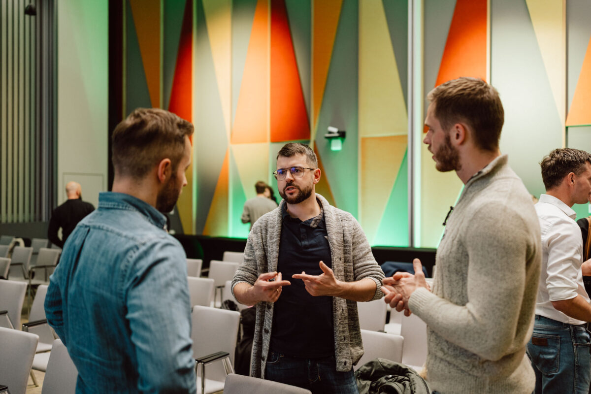 zdjecia eventowe aula polska bnp paribas warszawa 114 Trzech mężczyzn stoi w nowoczesnej sali konferencyjnej i prowadzi rozmowę. W tle widać kolorowe, geometryczne panele ścienne, a wokół nich kilka pustych krzeseł.