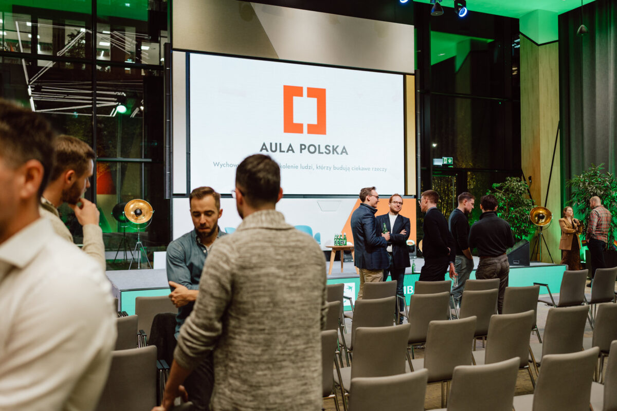 zdjecia eventowe aula polska bnp paribas warszawa 115 Grupa osób rozmawia przed dużym ekranem wyświetlającym logo "Aula Polska" w nowoczesnej przestrzeni eventowej z pustymi krzesłami i zielonym oświetleniem.