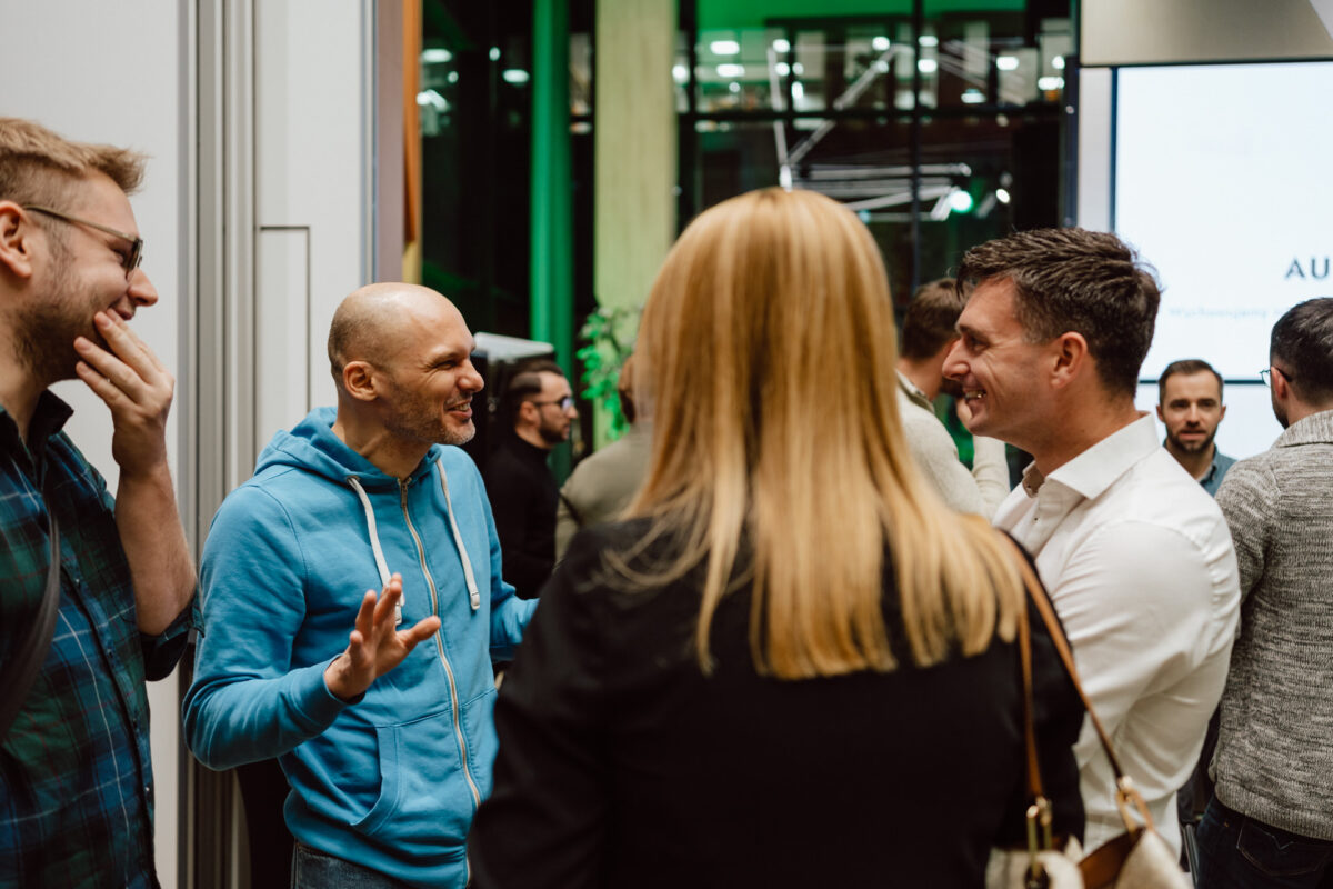 zdjecia eventowe aula polska bnp paribas warszawa 116 Grupa ludzi stoi w pomieszczeniu i prowadzi ożywioną rozmowę. Jeden mężczyzna w niebieskiej bluzie z kapturem gestykuluje podczas mówienia, inni uśmiechają się i słuchają. Otoczenie wydaje się być wydarzeniem towarzyskim lub networkingowym z dużymi oknami w tle.