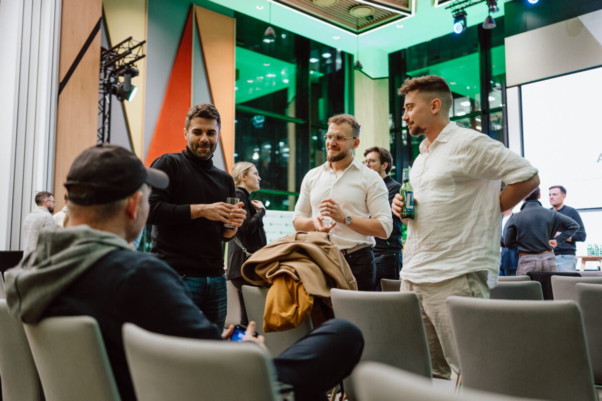 zdjecia eventowe aula polska bnp paribas warszawa 117 Grupa swobodnie ubranych osób stoi i rozmawia w nowoczesnej, jasno oświetlonej przestrzeni eventowej z geometrycznymi panelami ściennymi i dużymi oknami. Niektórzy trzymają drinki, podczas gdy inni siedzą, angażując się w rozmowę.