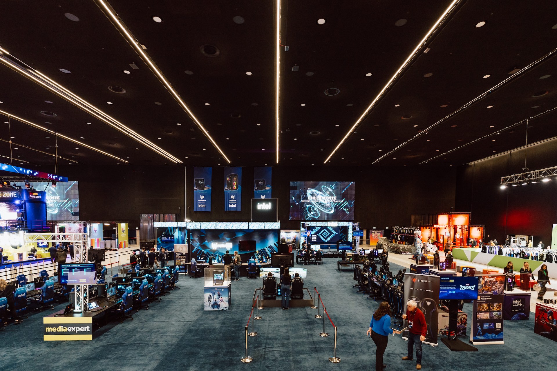 zdjecie stoiska expo IEM Katowice Intel Duża, jasno oświetlona hala kongresowa, w której odbywa się impreza gamingowa, z wieloma stoiskami, stanowiskami do gier, banerami i dużymi ekranami wyświetlającymi wizualizacje z gier - idealna do fotografii targowej, gdy uczestnicy spacerują i zwiedzają wystawy.