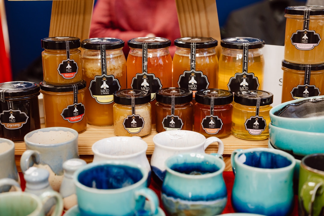 Auf dem Markt oder der Kunsthandwerksmesse stehen die Honiggläser auf Holzregalen hinter Reihen von handgefertigten Keramiktassen in verschiedenen Blau-, Weiß- und Brauntönen.