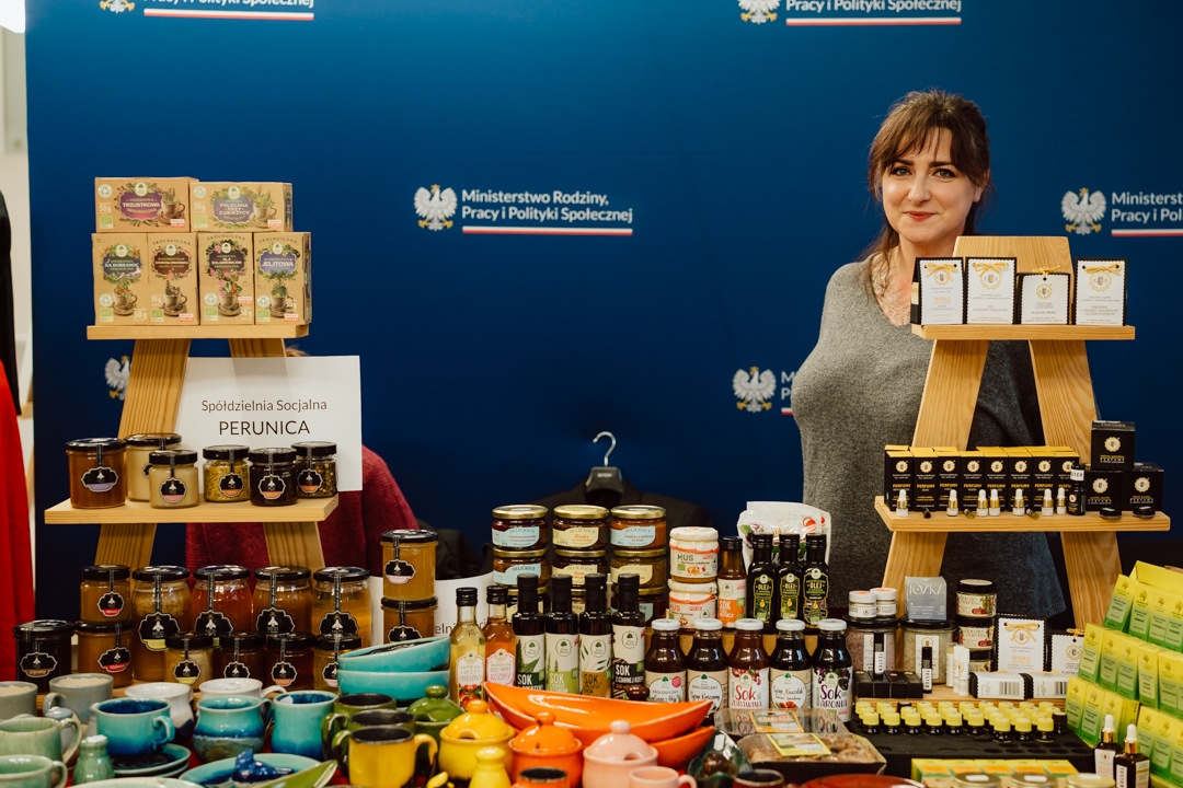 Eine Frau steht hinter einem Stand mit verschiedenen Gläsern, Flaschen, Tees und Keramikschalen. Auf blauem Hintergrund steht die Aufschrift 'Spółdzielnia Socjalna PERUNICA' mit dem Logo der polnischen Regierung.