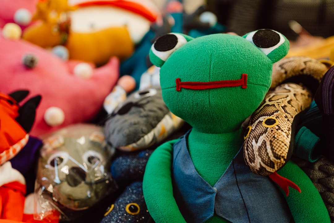 Ein grüner Plüschfrosch mit einer blauen Weste sitzt zwischen anderen Plüschtieren, darunter eine Schlange, ein Koala und verschiedene bunte Plüschtiere im Hintergrund.
