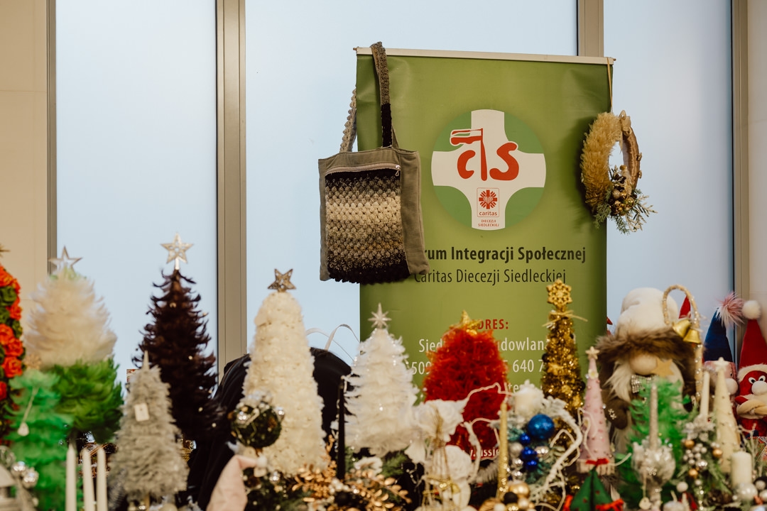 Eine Ausstellung von handgefertigtem Weihnachtsschmuck, darunter Weihnachtsbäume und Kränze, ist vor einem grünen Banner mit dem Logo und Namen des sozialen Integrationszentrums aufgebaut. Eine geflochtene Tasche und zwei Kränze hängen an dem Banner. 