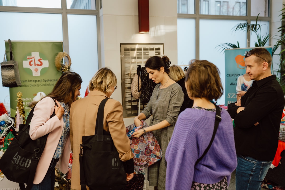 Eine Gruppe von Menschen steht um einen Tisch in einer Markthalle oder auf einem Jahrmarkt. Eine Frau in einem grauen Kleid zeigt der Gruppe bunte Stoffe. Im Hintergrund sind dekorative Gegenstände und Informationsbanner zu sehen.  