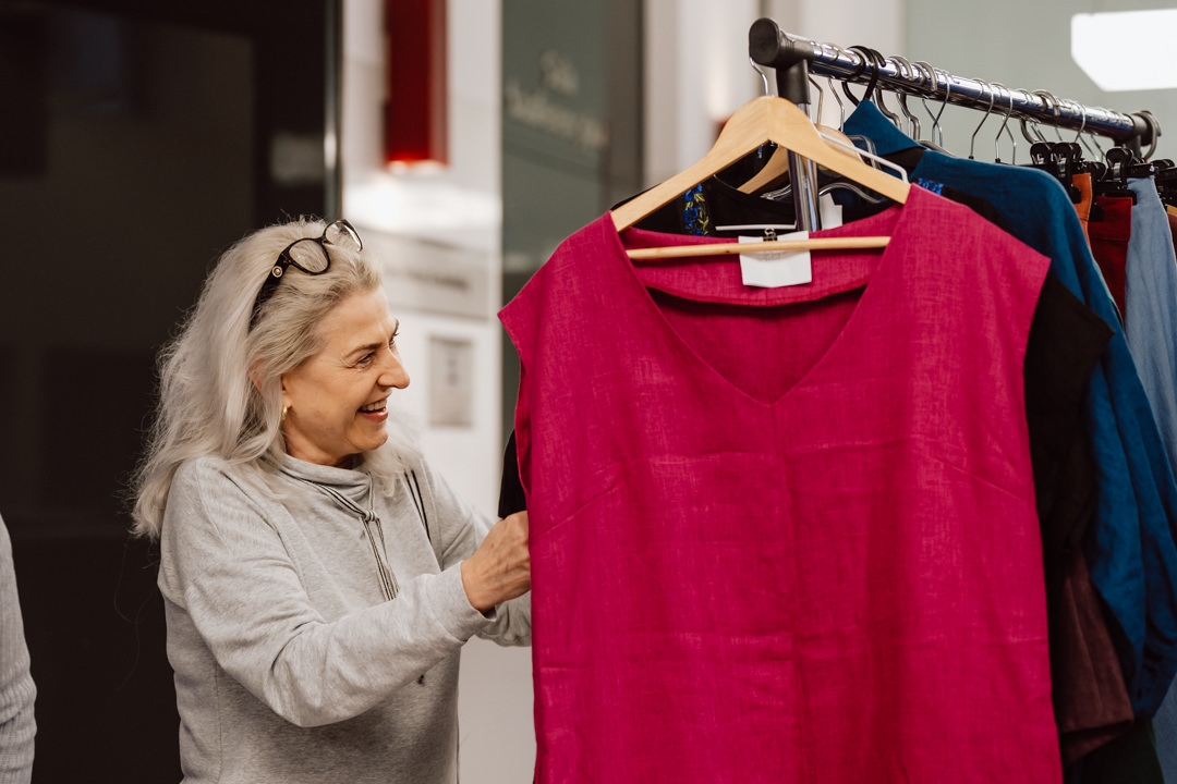Eine lächelnde ältere Frau mit grauen Haaren und einer Brille auf dem Kopf hält ein hellrosa Kleid hoch, während sie in einem Geschäft die Kleider auf einem Bügel betrachtet.