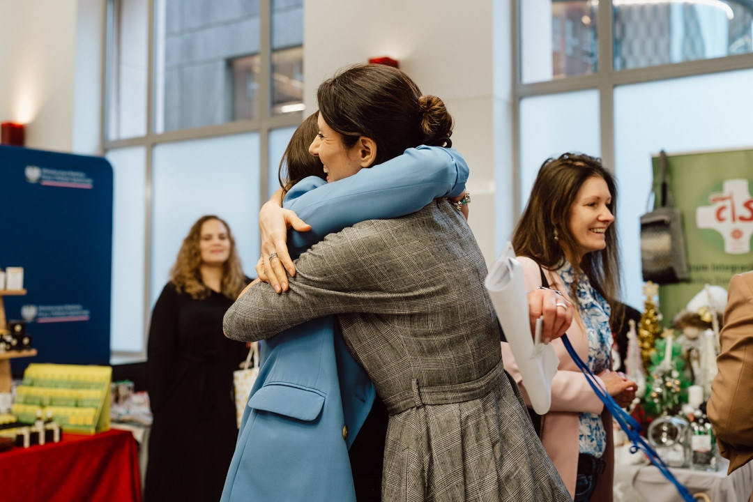 Zwei Frauen umarmen sich in einem überfüllten Innenraum, während andere Menschen herumstehen, einige lächelnd. Im Hintergrund stehen Tische mit verschiedenen Gegenständen und ein großes Fenster, das Tageslicht hereinlässt. 