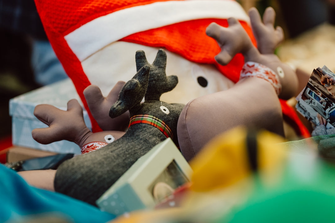 Eine Nahaufnahme der Weihnachtsdekoration, darunter ein Plüschrentier mit kariertem Schal und ein Kissen mit Weihnachtsmannmotiv, umgeben von eingepackten Geschenken und bunten Gegenständen.