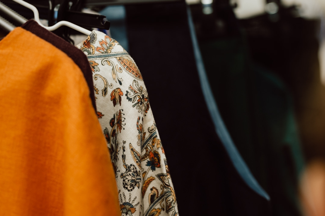 Nahaufnahme von Kleidungsstücken, die auf Kleiderbügeln hängen, darunter ein orangefarbenes Kleidungsstück, floral gemusterter Stoff und dunkle Stücke, die in einer Reihe ausgestellt sind.