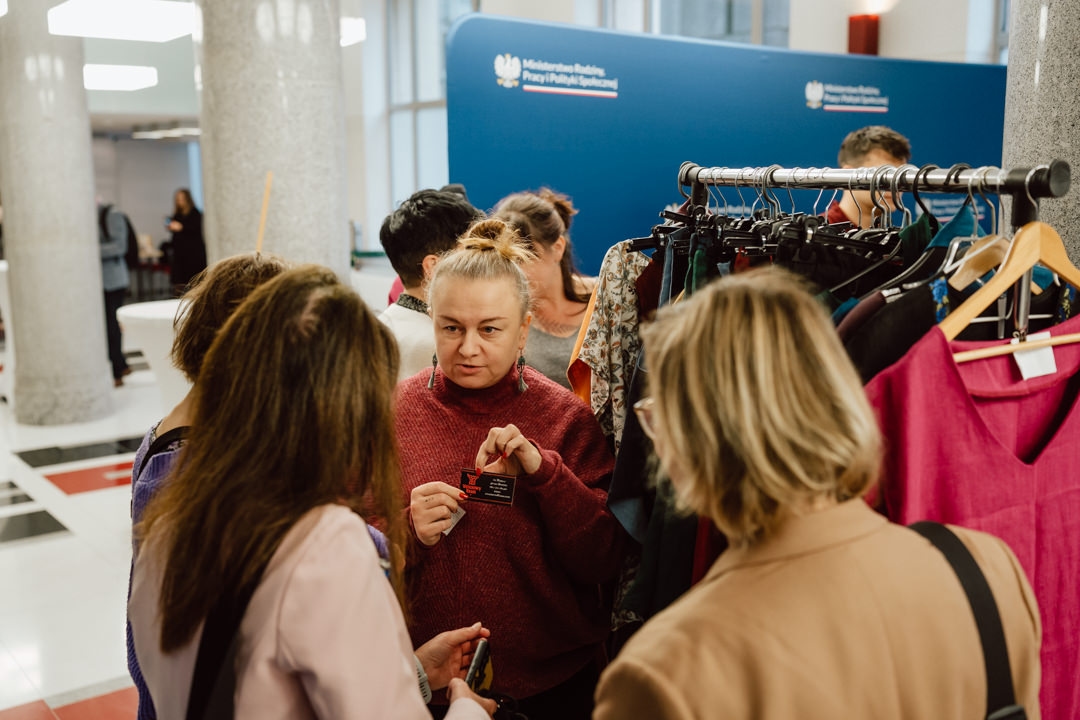 Eine Gruppe von Menschen steht in der Nähe eines Kleiderständers in einem Raum. Eine Person in einem roten Pullover hält eine Karte in der Hand und spricht mit den anderen. Im Hintergrund ist eine blaue Wand mit weißem Text und einem Logo zu sehen.  