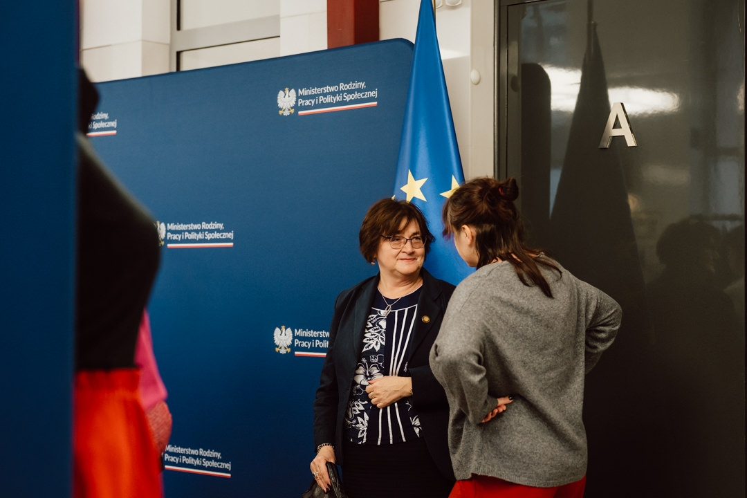 Zwei Frauen unterhalten sich vor einem blauen Hintergrund mit dem Logo des polnischen Ministeriums für Familienangelegenheiten und Sozialpolitik. Eine Flagge der Europäischen Union steht in der Nähe und eine dunkle Tür mit dem Buchstaben 'A' ist im Hintergrund zu sehen. 