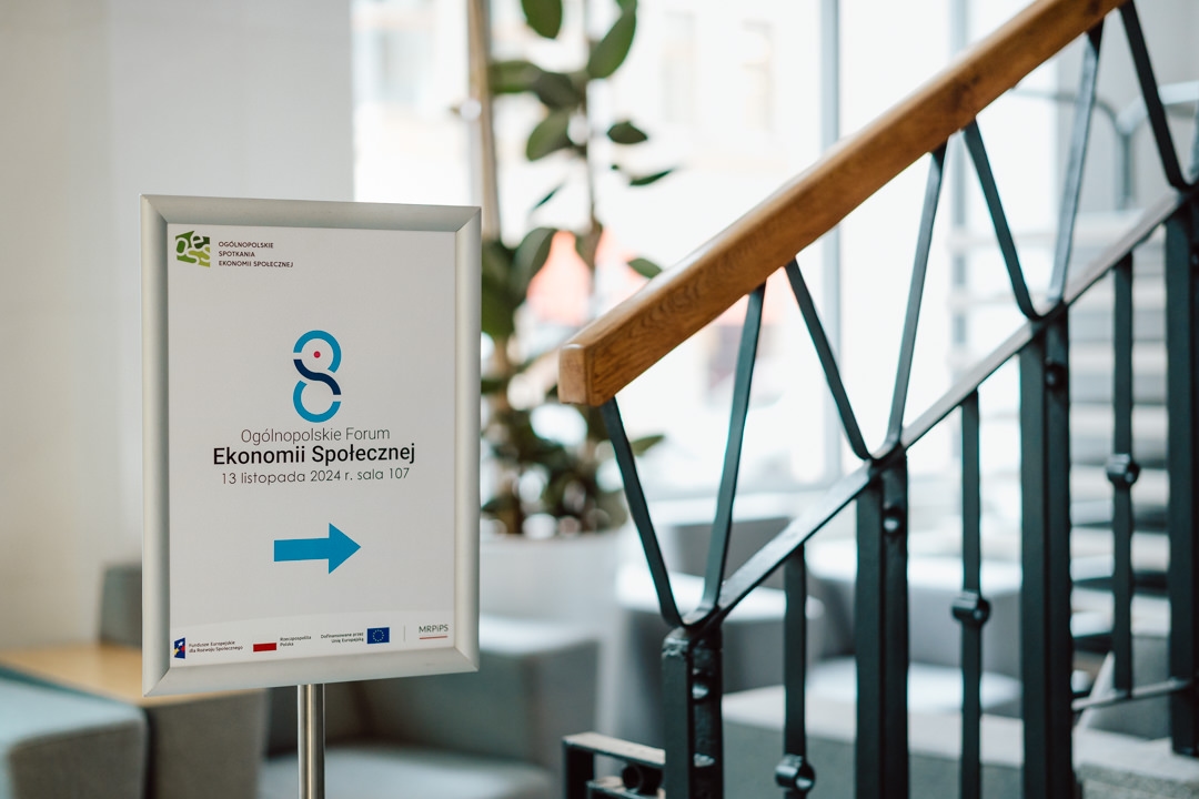 Ein Schild auf einem Ständer neben der Treppe weist in Richtung des Nationalen Forums für Sozialwirtschaft, das vom 1. bis 3. November 2024 in Raum 107 stattfinden wird. Der Pfeil auf dem Schild zeigt nach rechts.  