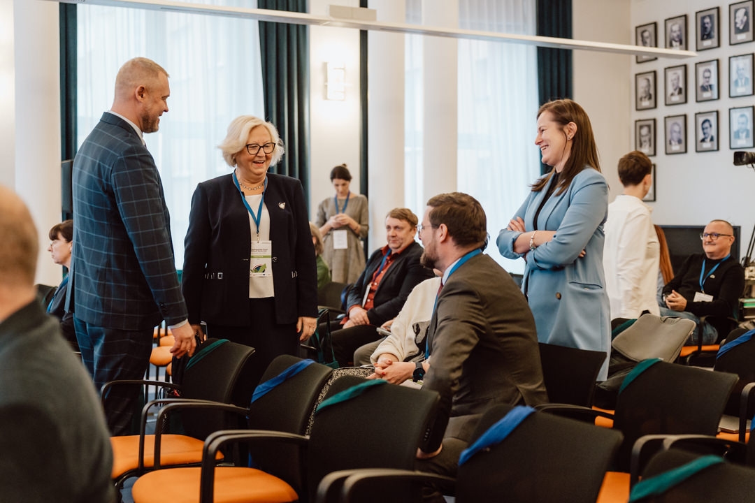 Eine Gruppe von Menschen in Geschäftskleidung plaudert und lächelt einander in einem Konferenzraum mit Reihen von orangefarbenen und schwarzen Stühlen an. Andere sitzen oder reden im Hintergrund und an den Wänden hängen gerahmte Porträts. 