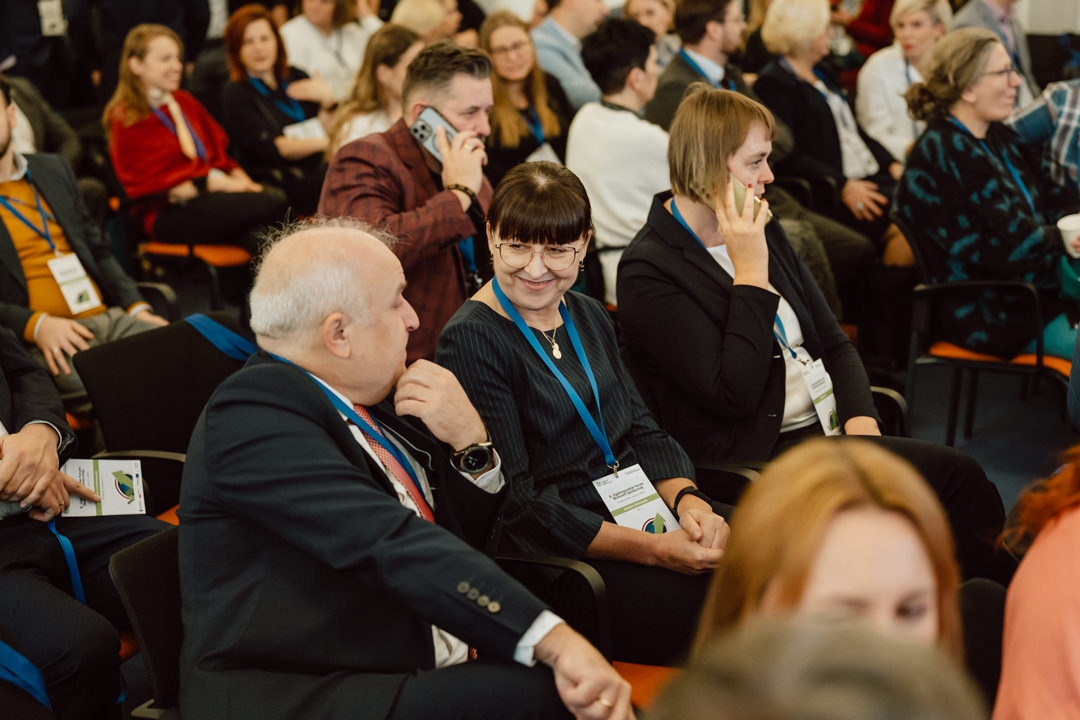 Eine Gruppe von Personen, von denen einige einen Ausweis tragen, sitzt bei einer Konferenz dicht beieinander. Die Frau in der Mitte lächelt und spricht mit dem Mann neben ihr, während andere mit ihren Telefonen beschäftigt sind oder sich unterhalten. 