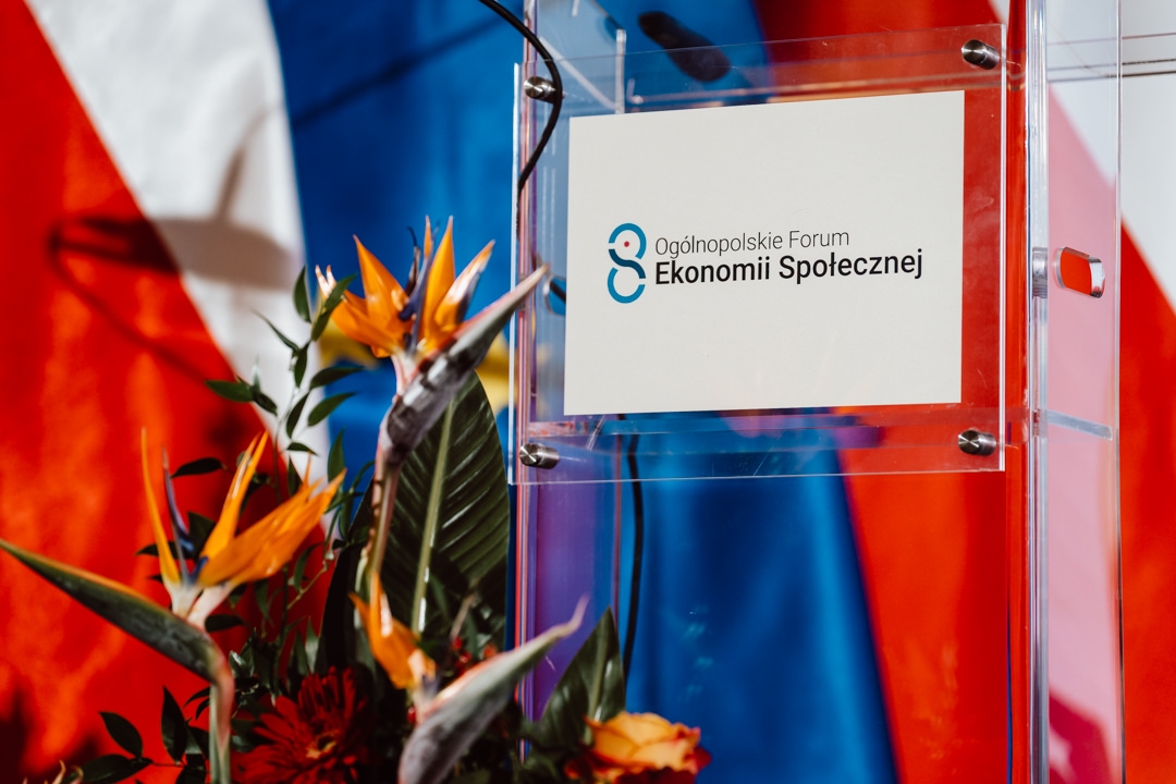 Ein transparentes Podium mit der Aufschrift 'Nationales Forum für Sozialwirtschaft' steht vor einem farbenfrohen Hintergrund aus Rot, Weiß und Blau und ist mit leuchtenden tropischen Blumen geschmückt.