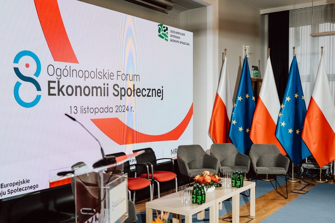 Konferenzraum mit einem großen Bildschirm, auf dem 'Nationales Forum der Sozialwirtschaft, 13. November 2024' angezeigt wird. Vier polnische und vier EU-Flaggen stehen hinter einer Reihe von leeren Stühlen, Tischen und Mikrofonen. 