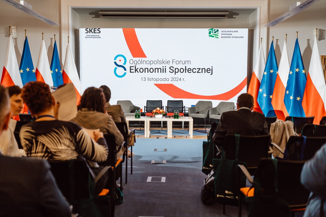 Konferenzraum mit Teilnehmern gegenüber der Bühne, mit zwei Stühlen und einem Tisch. Auf einem Bildschirm erscheint die Aufschrift 'Nationales Forum der Sozialwirtschaft, 13. November 2024'. An den Wänden sind polnische und EU-Flaggen angebracht.  