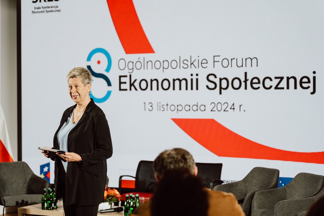 Eine Frau steht auf der Bühne und hält ein Tablet in der Hand. Sie spricht auf dem Nationalen Forum für Sozialwirtschaft. Der Titel und das Datum der Veranstaltung werden auf einem großen Bildschirm hinter ihr auf Polnisch angezeigt. 