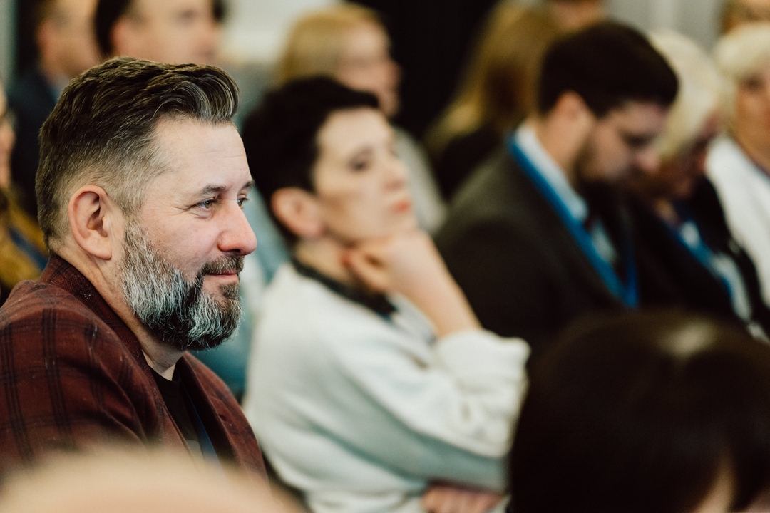 Eine Gruppe von Menschen sitzt dicht beieinander auf einer Party und hört aufmerksam zu. Ein bärtiger Mann in einer karierten Jacke steht im Mittelpunkt der Aufmerksamkeit, während andere leicht verschwommen im Hintergrund zu sehen sind. 