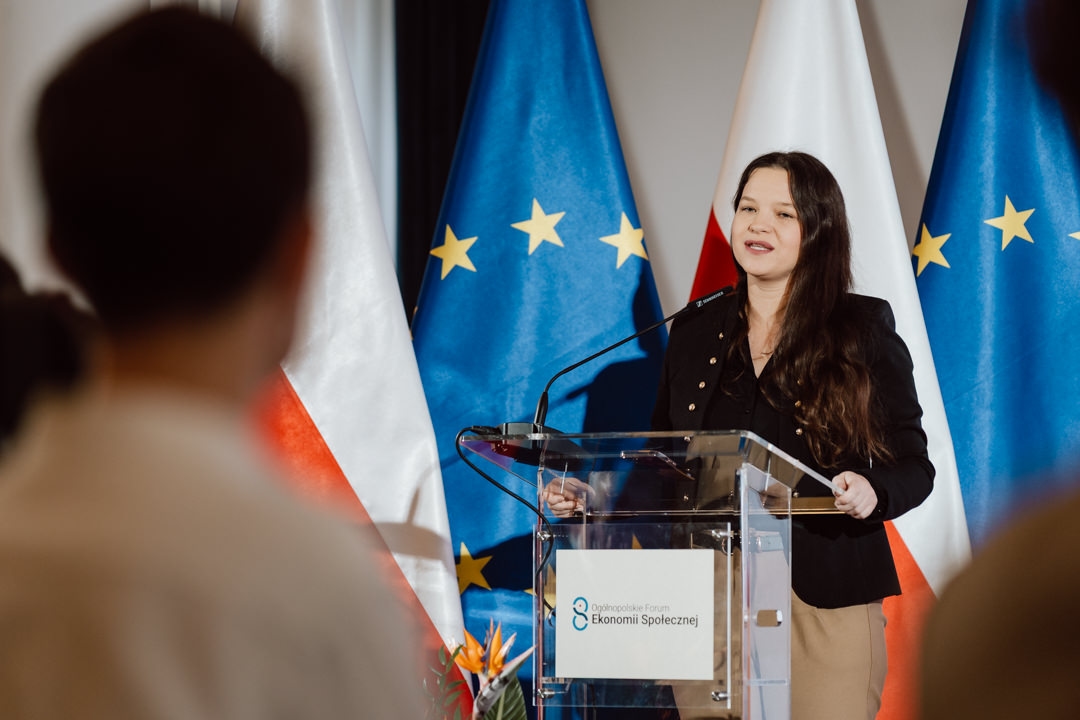 Eine Frau steht auf einem hellen Podium und wendet sich an das Publikum. Hinter ihr sind die Flaggen der Europäischen Union und Polens zu sehen. Auf dem Podium steht ein Schild mit polnischem Text. Die Szene findet in einem Raum statt, in dem im Vordergrund Menschen zuhören.   