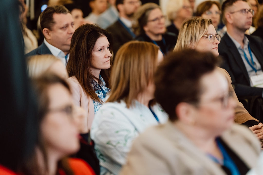 Eine Gruppe von Menschen sitzt dicht beieinander und hört einem Redner bei einer Veranstaltung oder Konferenz aufmerksam zu. Die meisten von ihnen tragen Geschäftskleidung und Ausweise, was auf ein professionelles Umfeld schließen lässt. 