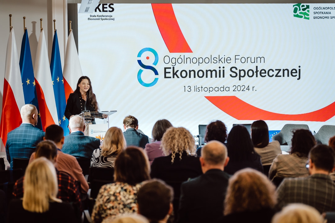 Eine Frau spricht auf dem Podium während des Nationalen Forums für Sozialwirtschaft am 13. November 2024. Die Teilnehmer sitzen vor einem Bildschirm, der den Namen der Veranstaltung anzeigt, mit Fahnen und Bannern im Hintergrund. 