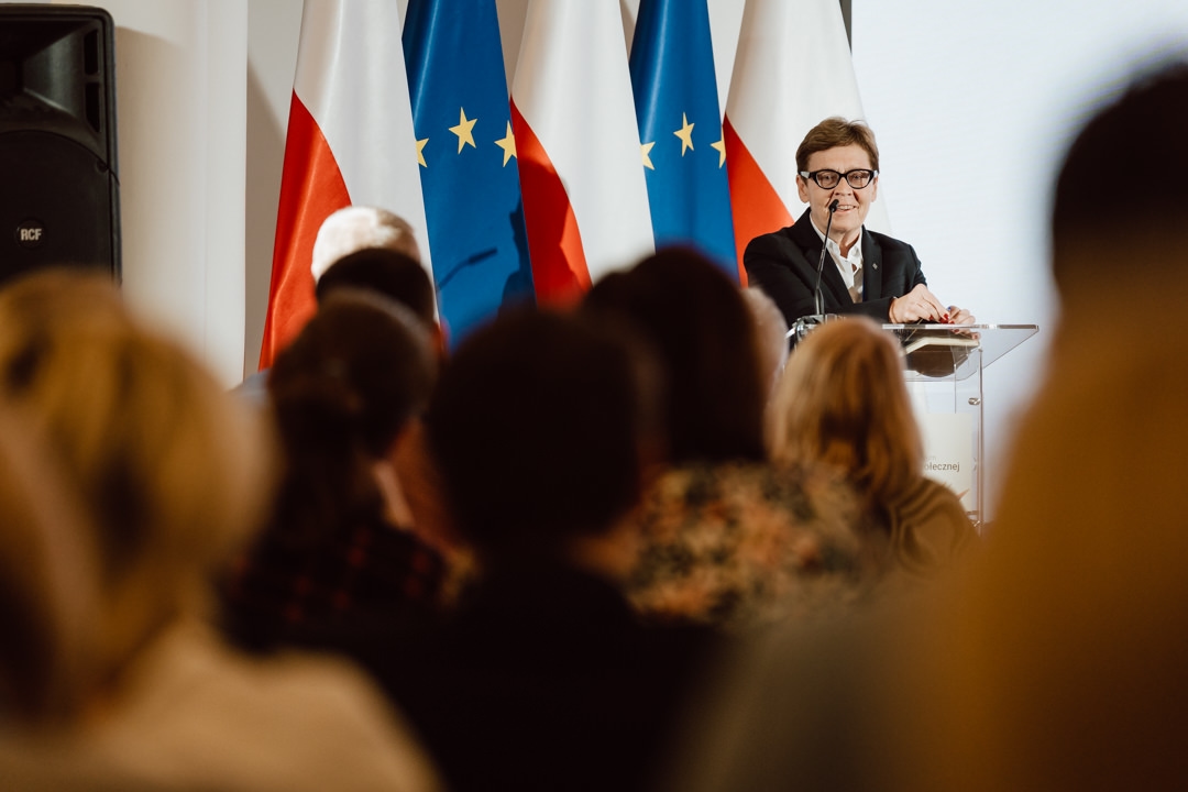 Eine Person im Anzug spricht auf einem Podium vor einem Publikum, während im Hintergrund die Flaggen Polens und der Europäischen Union zu sehen sind.