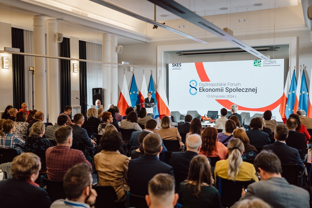Ein Konferenzraum voller Teilnehmer vor einer Bühne mit Rednern an einem Tisch und einem großen Bildschirm mit der Aufschrift "Opole Social Economy Forum, 13 November 2024" mit polnischen Flaggen im Hintergrund.