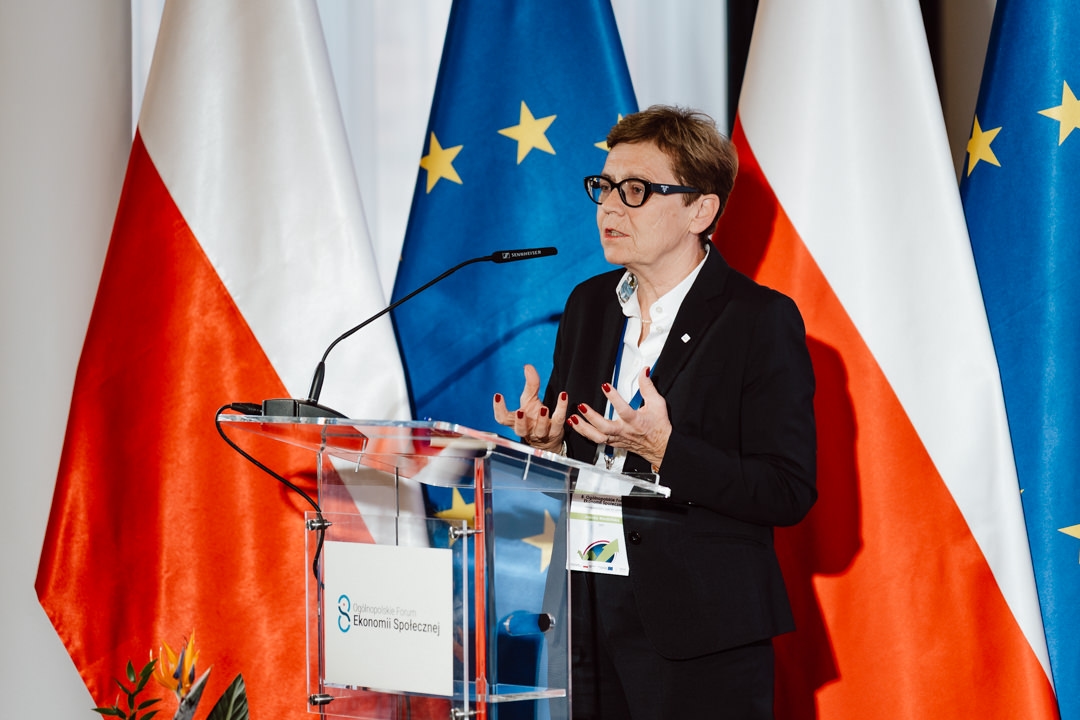 Eine Frau im dunklen Anzug und mit Brille spricht auf einem transparenten Podium mit Mikrofonen und gestikuliert mit ihren Händen. Hinter ihr sind die Flaggen von Polen und der Europäischen Union zu sehen. 