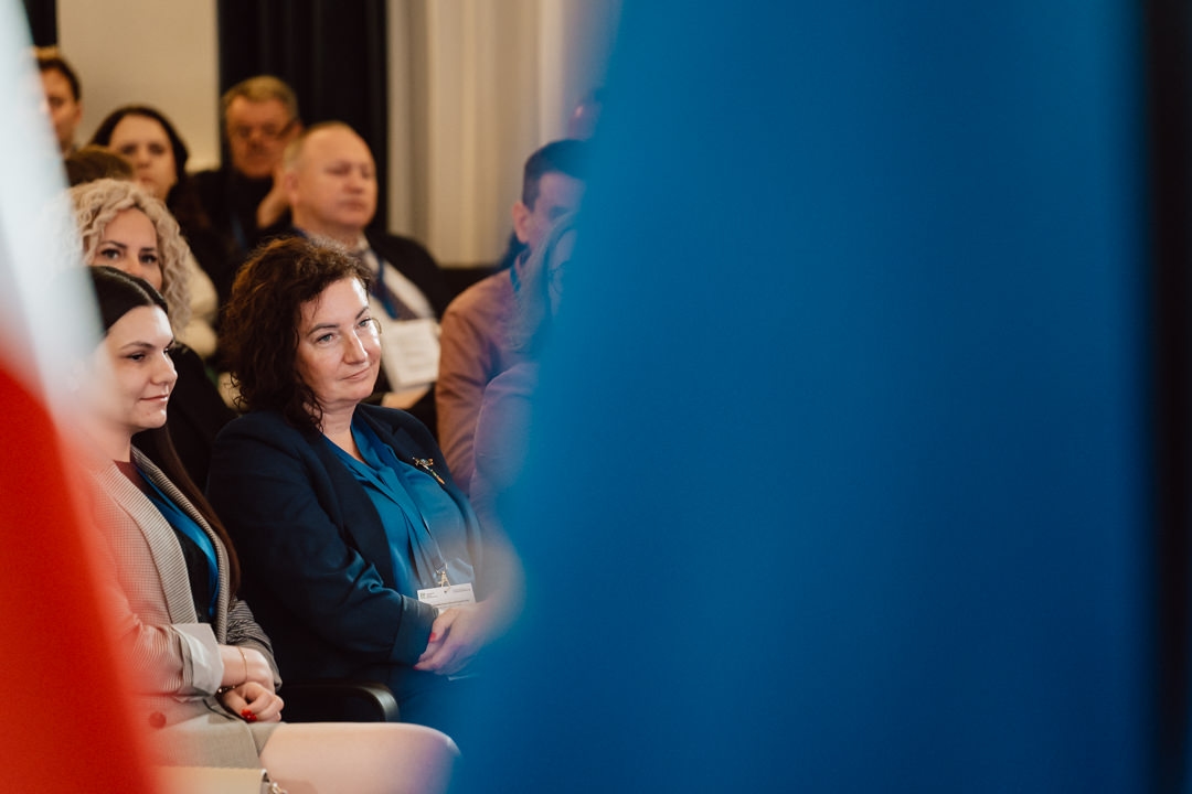 Eine Gruppe von Menschen sitzt aufmerksam im Publikum einer Indoor-Veranstaltung. Die Frau in der Mitte blickt nach vorne, während farbige Fahnen im Vordergrund verschwimmen.