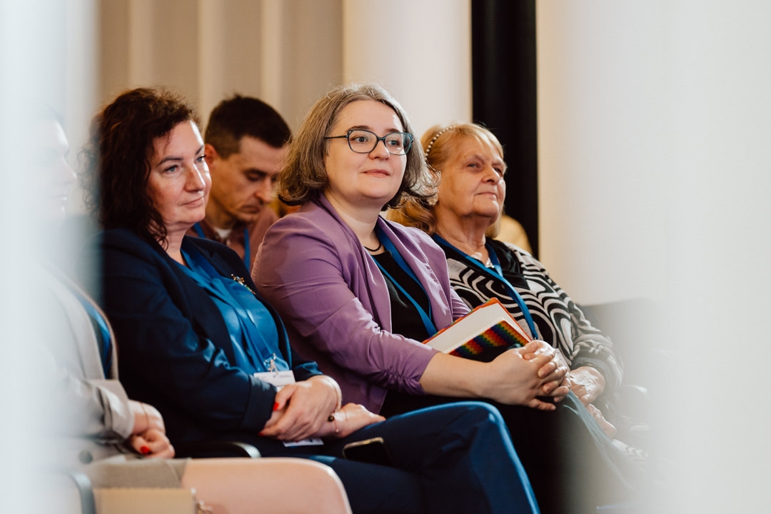 Mehrere Personen sitzen in einer Reihe auf etwas, das wie eine Konferenz oder ein Seminar aussieht, und hören aufmerksam zu. Die Frau in der Mitte, die einen Notizblock hält, trägt eine Brille und eine lila Jacke. Andere sitzen neben ihr, konzentriert auf das Geschehen.  
