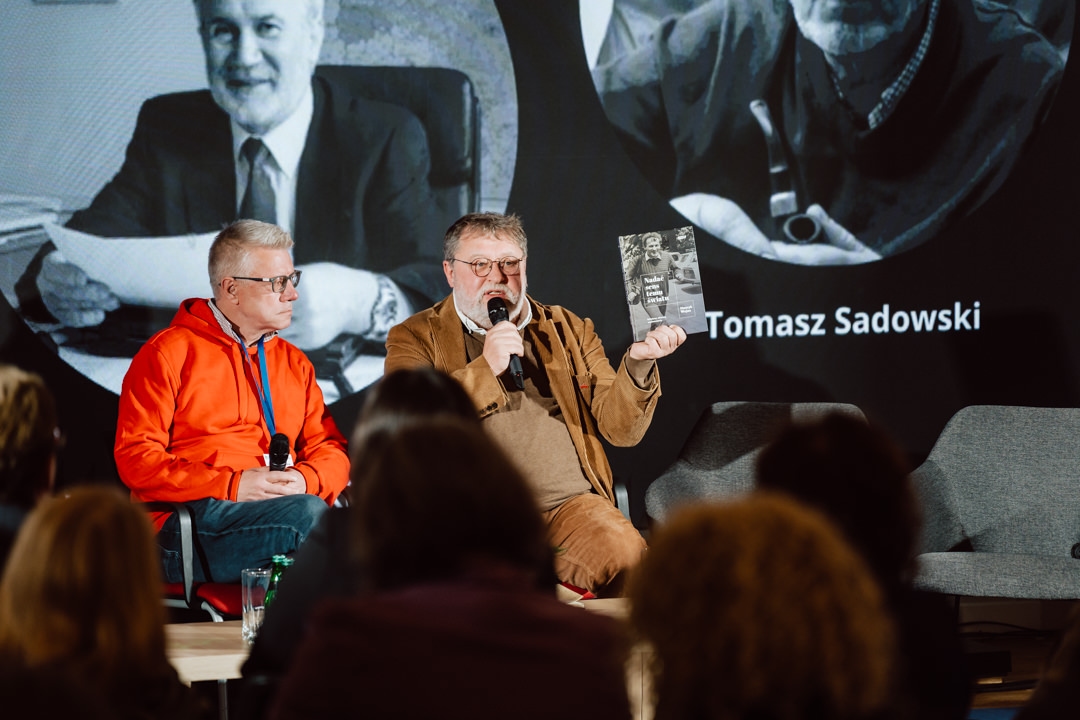 Zwei Männer sitzen während einer Podiumsdiskussion auf der Bühne. Einer von ihnen, der eine braune Jacke trägt, spricht in ein Mikrofon und hält ein Buch in der Hand. Der andere Mann in einem roten Sweatshirt hört zu. Das Publikum schaut zu, während Schwarz-Weiß-Porträts auf eine Leinwand projiziert werden.   