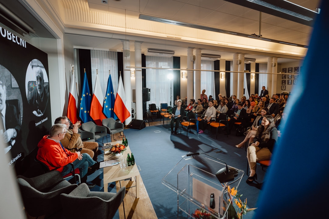 Eine Gruppe von Rednern sitzt auf einer Bühne vor einem Publikum in einem modernen, gut beleuchteten Raum, in dem polnische und EU-Flaggen hängen. Die Leute hören aufmerksam zu und an der Wand sind große schwarz-weiße Poster zu sehen. 