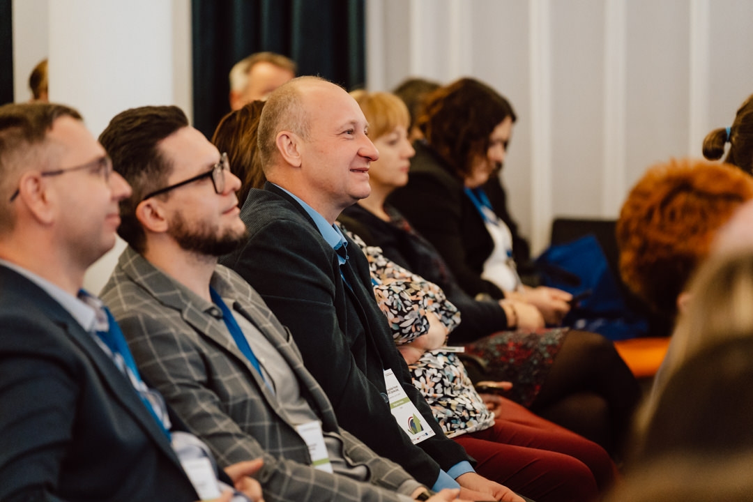 Eine Gruppe von Menschen, meist Männer in Geschäftskleidung, sitzt bei einer Veranstaltung in einer Halle dicht beieinander, hört aufmerksam zu und lächelt. Einige der Teilnehmer tragen Namensschilder. 