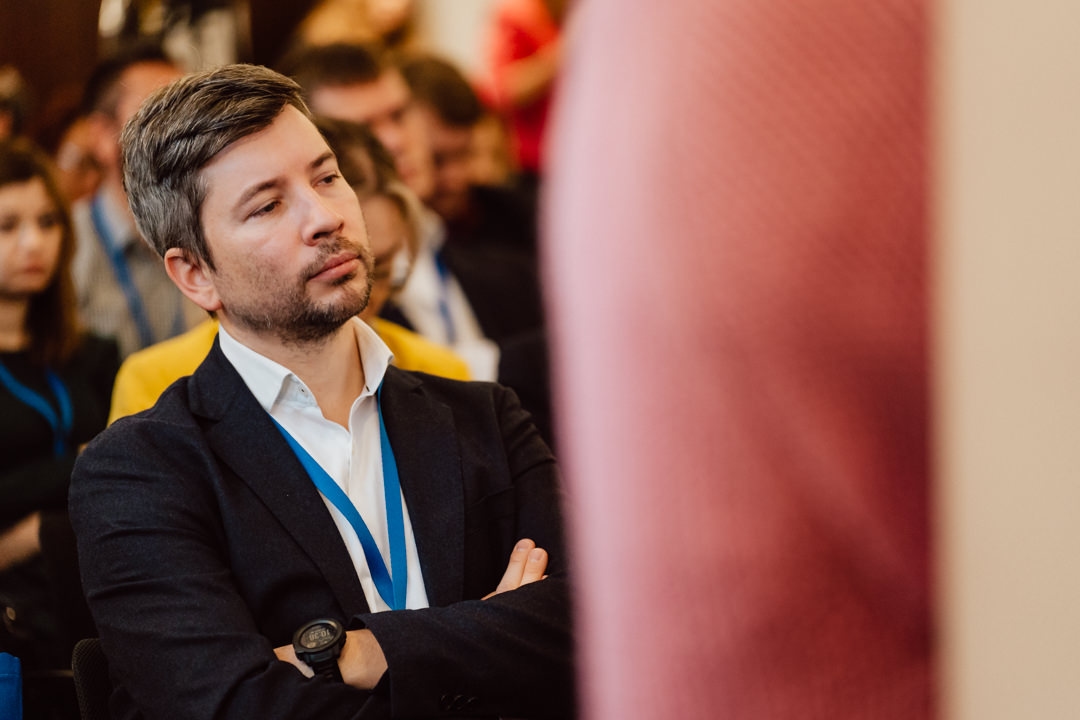 Ein Mann mit gestutztem Bart und dunkler Jacke sitzt mit verschränkten Armen da und hört aufmerksam einer Veranstaltung im Raum zu. Im Hintergrund sitzen Menschen, und im Vordergrund ist teilweise ein rosafarbener Lautsprecher zu sehen. 