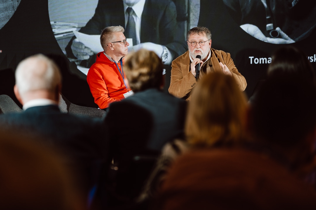 Zwei Männer sitzen auf der Bühne und diskutieren, einer hält das Mikrofon und spricht, während der andere zuhört. Die Zuschauer sitzen im Vordergrund, mit Blick auf die Redner. Hinter ihnen befindet sich ein großer Bildschirm mit Bildern und Text.  