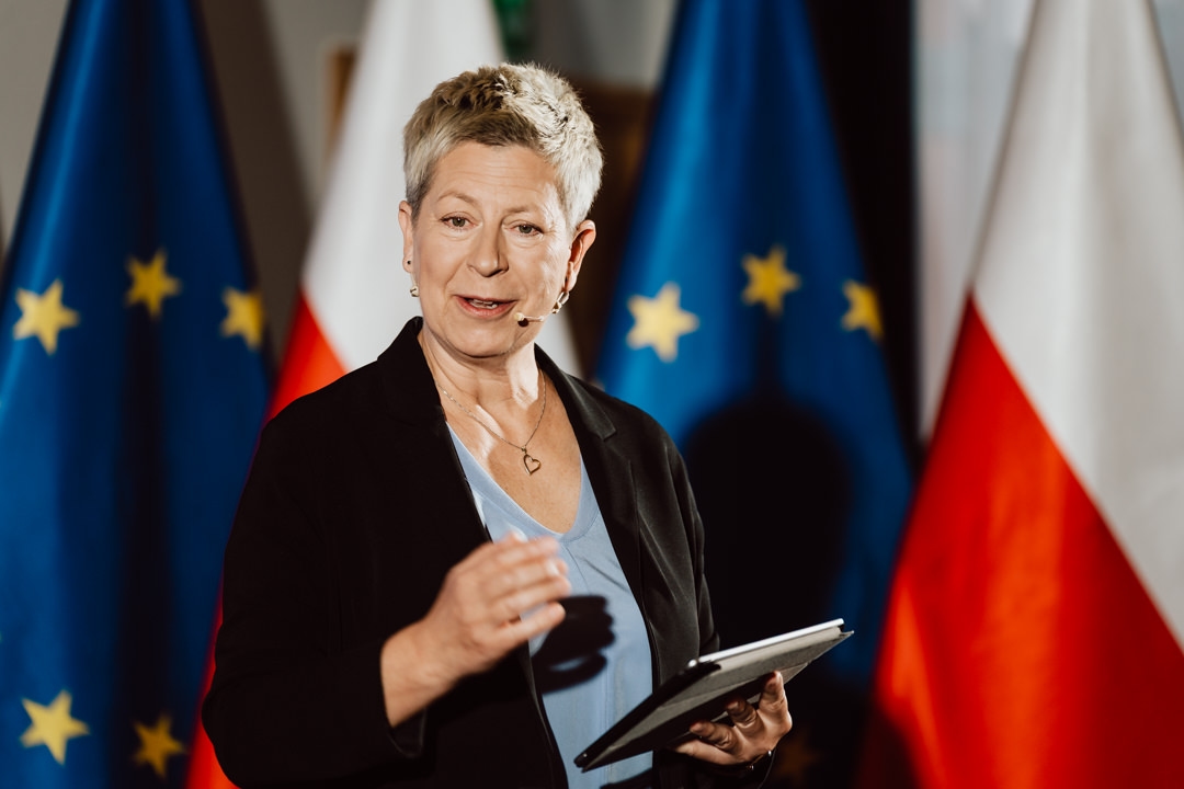 Eine Frau mit kurzen blonden Haaren spricht mit einem Tablet in der Hand, während sie vor den Flaggen der Europäischen Union und Polens steht. Sie trägt eine schwarze Jacke und ein hellblaues Oberteil und benutzt ein Headset-Mikrofon. 