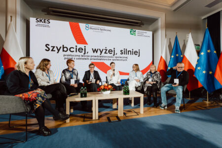 Fotograf na forum ekonomii spolecznej 042 Panel siedmiu osób siedzi na scenie przed dużym ekranem z polskim tekstem na konferencji. W pobliżu stoją trzy flagi narodowe. Paneliści są zaangażowani w dyskusję, niektórzy trzymają mikrofony.